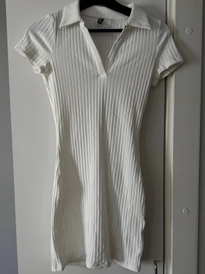 Vit ribbad kortklänning från H&M - Vit ribbad kortklänning från H&M med krage och v-ringning. Klänningen har korta ärmar och är tillverkad i ett stretchigt material som sitter snyggt på kroppen. 