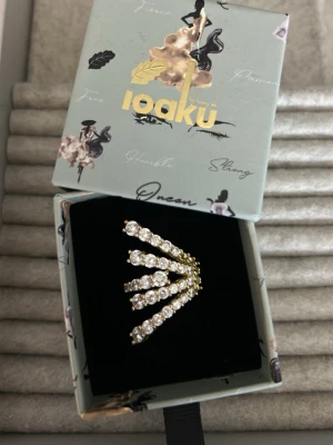 Guldfärgad statementring från Ioaku - Unik guldfärgad ring från Ioaku med fem utsvängda rader dekorerade med gnistrande klara stenar. Ringen har en modern och iögonfallande design som verkligen sticker ut. Perfekt accessoar för dig som gillar att synas och vill ha något extra.