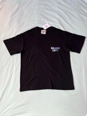 Svart t-shirt från Gallery Dept. - Svart t-shirt från Gallery Dept. med tryckt logga i vitt och blått på bröstet. Klassisk rund hals och korta ärmar. Tillverkad i mjuk bomull för en skön och avslappnad stil.