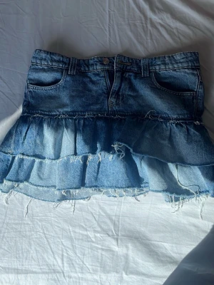 Jeanskjol med volanger från Gina Tricot - Snygg jeanskjol från Gina Tricot i ljusblå denim med volangdetaljer och råa kanter. Kjolen har bälteshällor och en kort passform som ger en trendig look. Perfekt för dig som gillar en avslappnad och cool stil.