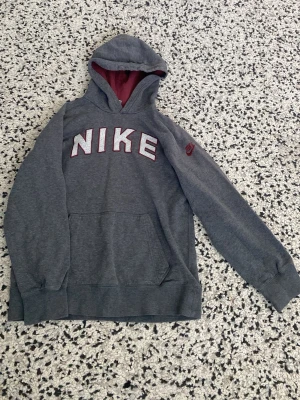 Grå Nike hoodie med röd detalj - Snygg grå hoodie från Nike med stor vit och röd logga på bröstet och röd Nike-detalj på ärmen. Hoodien har magficka och röd insida i huvan. Perfekt för en avslappnad och sportig stil.