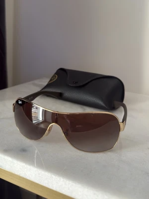 Ray-Ban solglasögon  -  Ray-Ban solglasögon med shield-formade bruna glas och guldfärgade detaljer. Bågarna är svarta och glasen har en lätt toning, Köpta i Italien 