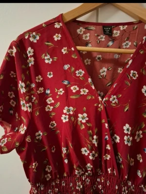 Röd blommig midiklänning från Shein - Röd klänning från Shein med vitt och blått blommönster, v-ringad hals och korta ärmar. Klänningen har resår i midjan för en bekväm passform och är gjord i ett mjukt, lätt material. Perfekt för sommarens alla tillfällen.