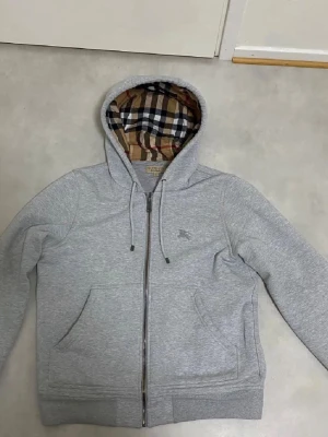 Grå tröja från Burberry - Säljer en stilren grå jacka från Burberry med klassiskt rutigt mönster på insidan. Jackan har en mjuk och bekväm känsla, perfekt för kyligare dagar. Den har en dragkedja framtill och en huva för extra skydd mot väder och vind.