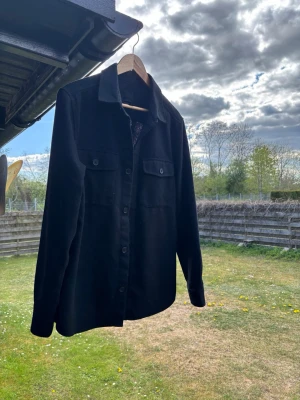 Svart overshirt med fickor - Stilren svart overshirt med klassisk krage och knäppning framtill. Två bröstfickor med lock och knappdetaljer ger en snygg look. Långa ärmar med manschett och knapp. Perfekt lager på lager-plagg för en trendig stil.