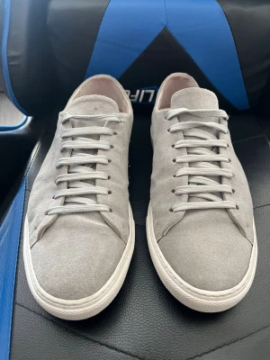 Ljusgråa sneakers i mocka - Snygga ljusgrå sneakers från William Stouch i mjuk mocka med klassisk låg profil och vita sulor. Skorna har matchande grå skosnören och en stilren design som passar till det mesta. Perfekta för dig som gillar minimalistisk och clean stil.