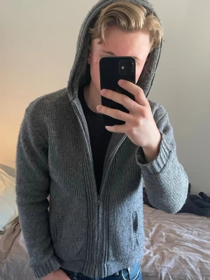 Grå stickad cardigan med luva - Grå stickad cardiganjacka med luva och dragkedja framtill. Jackan har två fickor och ribbade muddar vid ärmslut och nederkant. Insidan av luvan är fodrad med mjuk päls för extra komfort. Perfekt för kyliga sommarkvällar och enkel att matcha.