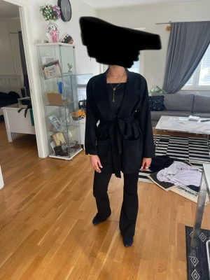Svart kappa med knytskärp - Stilren svart kappa med knytskärp i midjan och klassisk krage. Lång modell med raka ärmar och utan synliga knappar. Perfekt för dig som gillar en minimalistisk look. Materialet ser ut att vara mjukt och följsamt.