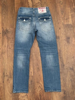 True Religion Jeans ”Joey SuperT” - Säljer ett par klassiska blå ”Joey SuperT” jeans från True Religion med raka ben och snygga vita kontrastsömmar. Jeansen har två bakfickor med lock och vita knappar samt en patch med logga bak i midjan. Perfekta för en avslappnad och trendig look.
