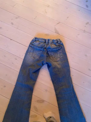 Blå bootcut jeans med fickdetaljer - Ett par klassiska blå bootcut jeans med snygga fickdetaljer bak och lätt slitning framtill. Jeansen har normalhög midja och är tillverkade i denim med en skön passform. Perfekta för en avslappnad och trendig look. I storlek 164 och priset kan diskuteras