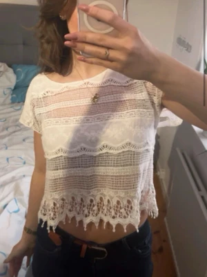 Vit spetsig croppad blus - Superfin vit croppad blus med spetsdetaljer och korta ärmar. Toppen har ett vackert virkat mönster och dekorativ kant nertill. Perfekt att styla med höga jeans eller kjol för en trendig look.