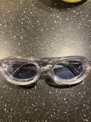 Transparenta solglasögon från Arigato - Snygga solglasögon från Arigato med transparent båge och svarta glas. Glasögonen har en rundad form och ger en cool vibe till din outfit. Perfekta för dig som vill sticka ut med en unik accessoar.