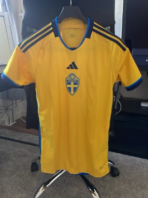 Sverige fotbollströja Adidas gul - Gul fotbollströja från Adidas med Sveriges landslagsemblem på bröstet och blå detaljer vid krage och ärmslut. Tröjan har svarta Adidas-ränder på axlarna och texten 'Sverige' i nacken. Perfekt för match eller träning.