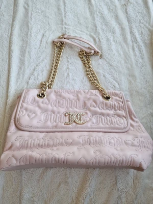 Ljusrosa Juicy Couture handväska - Ljusrosa handväska från Juicy Couture med quiltat mönster och logotyp broderad över hela väskan. Väskan har guldfärgade kedjehandtag och ett dekorativt JC-spänne framtill. Tillverkad i konstläder med mjuk finish och hjärtdetaljer.