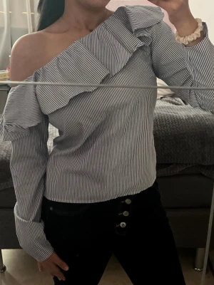 Randig offshoulder-blus med volang - Snygg blus med smala blå och vita ränder, offshoulder-modell och volangdetalj längs halsringningen. 