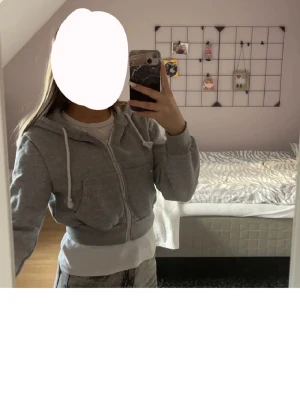 Grå croppad hoodie med dragkedja - Snygg grå croppad hoodie med dragkedja framtill och stora fickor. Har huva med vita dragsnören och breda muddar vid ärmslut och midja. Perfekt för en chill och avslappnad stil. Mjukt, skönt och tjock tyg. Passar s/m