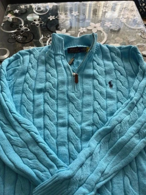 Ljusblå kabelstickad tröja Polo Ralph Lauren - Ljusblå kabelstickad tröja från Polo Ralph Lauren med halv dragkedja och broderad logga på bröstet. Tröjan har ribbade muddar och klassisk passform. Perfekt för dig som gillar stilrena och bekväma plagg.