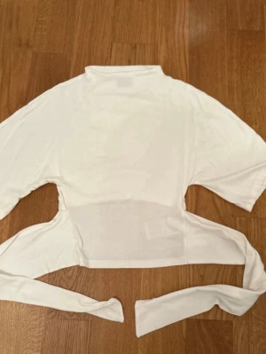 Vit croppad topp med knyt - Trendig vit croppad topp med knyt i midjan och korta ärmar. Toppen har en hög hals och är tillverkad i ett tunt, mjukt material som ger en snygg och luftig känsla. Perfekt att styla med jeans eller kjol för en cool look.