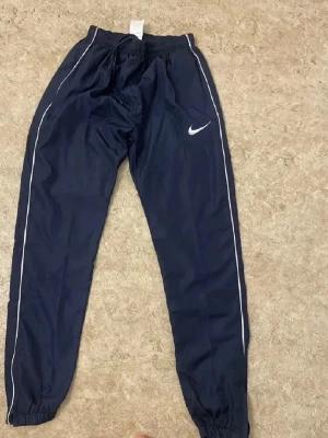 Mörkblå Nike mjukisbyxor med vita detaljer - Mörkblå mjukisbyxor från Nike med vita ränder längs benen och klassisk Nike-logga på låret. Byxorna har resår i midjan och vid bensluten för en skön passform. Perfekta för chill eller träning.
