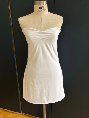 Vit kort klänning - Snygg vit axelbandslös klänning med lätt rynk framtill vid bysten. Avtagbart halterneckband med tryckknappar (se bild 6-7) Klänningen är kort och har en enkel, rak passform som ger en stilren look. Perfekt för dig som gillar minimalistisk stil och vill ha något bekvämt och luftigt. Tyget är stretchigt, nästan som baddräktstyg.