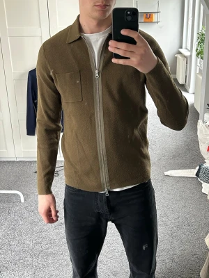 Olivgrön overshirt med dragkedja - Stilren olivgrön overshirt med rak passform och hel dragkedja framtill. Från Samsoe Samsoe. Storlek S.