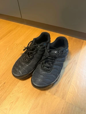 Nike Air Max Plus Tn helsvarta sneakers - Nike Air Max Plus Tn sneakers i helsvart med klassisk vågig design och Tn-logo på hälen. Ovandel i mesh och syntet, robust sula med synlig Air Max-dämpning. Perfekta för dig som gillar streetwear och sportig stil.