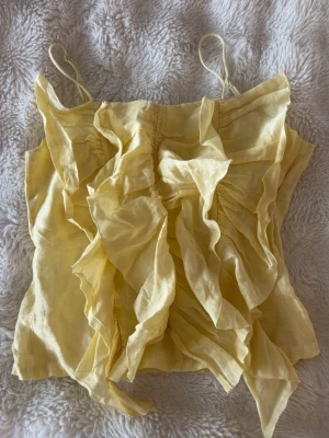 Gul volangtop  - Söt gul topp från H&M med tunna axelband och stora volanger framtill. Tillverkad i ett lätt och luftigt tyg som ger en somrig känsla. Perfekt för dig som gillar statement-plagg och vill sticka ut lite extra.