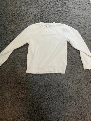 Vit basic sweatshirt med rund hals - En stilren vit sweatshirt med rund halsringning och långa ärmar. Tröjan har ribbade muddar vid ärmslut och nederkant, perfekt för en clean och enkel look. Tillverkad i mjukt material som känns skönt mot huden.