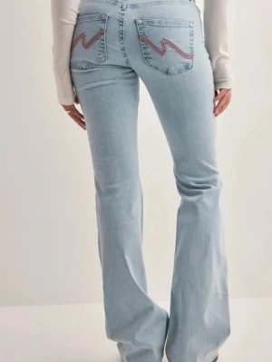 Ljusblå bootcut jeans med brodyr - Säljer ett par ljusblå bootcut jeans med snygg röd och blå brodyr på bakfickorna. Jeansen har klassisk femficksdesign och är tillverkade i denim med lätt stretch. Perfekta för en trendig och avslappnad look.