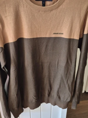 Beige och brun stickad tröja Armani Jeans - Stickad tröja från Armani Jeans i tvåfärgad design med beige upptill och mörkbrun nedtill. Rund halsringning och diskret logga på bröstet. Perfekt för dig som gillar stilrena och klassiska plagg med en modern touch.