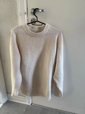 Beige stickad tröja med rund hals - En stilren beige stickad tröja med rund halsringning och ribbade muddar vid ärmslut och nederkant. Tröjan har en rak passform och är perfekt för lager-på-lager. Materialet känns mjukt och mysigt, vilket gör den skön att bära under kyliga dagar.