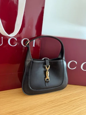  Gucci Jackie 1961 mini - Snygg svart Gucci Jackie 1961-kollektionen i slätt läder med guldfärgad metalldetalj framtill. Väskan har en klassisk, avlång form och kort axelrem. Följer även med ett långt axelrem. Perfekt för dig som gillar lyxiga accessoarer med ikonisk design. Höjd 13cm bredd 19 cm bredd 3,5cm