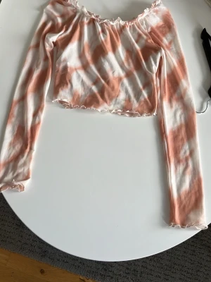 Offshoulder croppad topp i rosa och vit - Trendig offshoulder topp med långa ärmar i croppad modell. Toppens tyg har ett batikliknande mönster i ljusrosa och vitt, med volangkant vid halsringning och ärmslut. Perfekt för dig som gillar en söt och avslappnad stil.