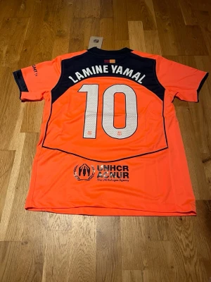 Barcelona third kit lamine yamal #10 - Snygg orange FC Barcelona matchtröja med Lamine Yamal och nummer 10 på ryggen. Tröjan har mörkblå detaljer, klubbmärke och Nike-logga framtill samt UNHCR-tryck bak. Tillverkad i lätt och ventilerande material, perfekt för fotbollsträning eller match.