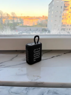 JBL GO 3 bärbar högtalare svart - Snygg och kompakt JBL GO 3 bärbar högtalare i svart med robust tygmaterial och praktisk ögla för enkel upphängning. Perfekt för att ta med sig överallt och spela musik trådlöst via Bluetooth. Stilren design med tydliga knappar på ovansidan.