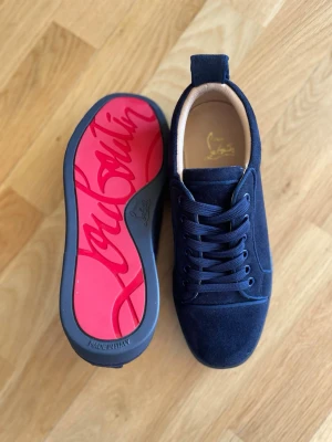  Christian Louboutin  - Snygga mörkblå sneakers från Christian Louboutin i mocka med klassisk röd sula och diskret logga. Skorna har rund tå, svarta snören och låg profil. Perfekta för dig som vill sticka ut med exklusiv stil och ikonisk design.