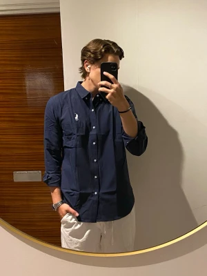 Skjorta från Ralph Lauren - Snygg mörkblå skjorta från Ralph Lauren med klassisk krage och vita knappar. Skjortan har lång ärm och ikonisk broderad logga på bröstet. Perfekt för en stilren look och enkel att matcha med olika outfits. Storlek M. Jag är 177 cm.