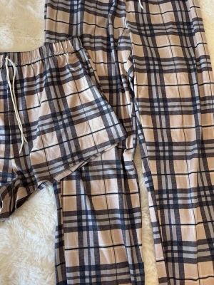 Rutiga pyjamasbyxor och shorts - Snyggt set med rutiga pyjamasbyxor och matchande shorts i beige och svart. Båda har resår i midjan och snörning för extra komfort. Perfekt för chill dagar hemma. Mjukt och lätt material som känns skönt mot huden.