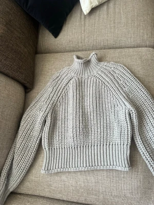 Ljusgrå stickad cropped tröja - Säljer en ljusgrå stickad tröja med hög hals och bred ribbad mudd nertill. Tröjan är cropped och har långa ärmar med grovstickat mönster. Perfekt för lager-på-lager och chill dagar.