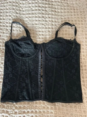 Svart korsettopp i spets  - Svart korsettopp med spets och justerbara axelband. Framsidan har bygel och vacker spets, baksidan har snörning. Materialet är spets, vilket ger en lyxig och feminin känsla. Perfekt för att skapa en statement-look. Använd 1 gång