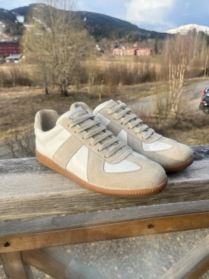 Vita Maison margielas  - Snygga sneakers i beige mocka och vitt läder med klassisk gummisula. Modellen har lågt skaft och stilrena linjer, perfekt för en clean och trendig look. Skorna har matchande beige snörning och diskret branding på plösen. Perfekt nu inför sommaren ☀️