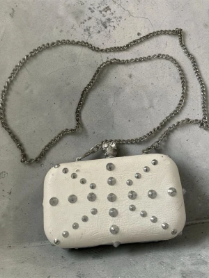 Vit kuvertväska med nitar och dödskalle - Cool vit kuvertväska med silverfärgade nitar i mönster på framsidan och en unik dödskalle-detalj på låset. Väskan har en lång silverkedja och är tillverkad i konstläder med en hård, boxig form. Perfekt för dig som gillar edgy accessoarer.