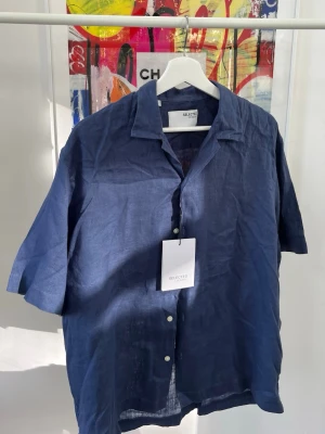 Blå linneskjorta från Selected Homme - Snygg blå skjorta i linne från Selected Homme med kort ärm och klassisk krage. Skjortan har knappar framtill och en avslappnad passform, perfekt för varma dagar. Den är något stor i storleken, lite oversized-känsla. 