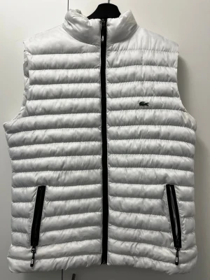 Vit dunväst från Lacoste - Snygg vit dunväst från Lacoste med ståkrage och svarta dragkedjor framtill och på fickorna. Västen har det klassiska Lacoste-märket på bröstet. Köpte den utomlands pga kalla vädret men har inget behov. Den är liten i storlek passar L. Om du intresserad av byta hör av dig.