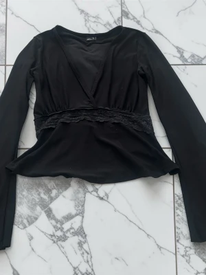 Svart omlottopp med spetsdetalj - Svart långärmad omlottopp från Gina Tricot med spetsdetalj under bysten. Toppen har en figurnära passform och är tillverkad i mjuk polyesterblandning med elastan för extra stretch. Perfekt för dig som gillar stilrena och trendiga plagg.