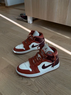 Air Jordan 1 Mid röd och vit - Snygga Air Jordan 1 Mid sneakers i rött och vitt läder med klassisk svart swoosh på sidan. Skorna har högt skaft, perforerad tåbox och röda skosnören. Perfekta för dig som gillar streetwear och vill sticka ut med en ikonisk modell.