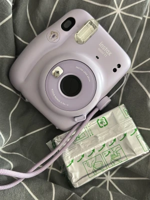 Lila Instax Mini 11 kamera - Säljer en lila Fujifilm Instax Mini 11 direktfilmskamera. Kameran har en rund, kompakt design i plast och levereras med handledsrem. Perfekt för att ta instant-bilder med vänner och få foton direkt i handen.