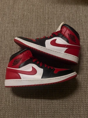 Air Jordan 1 Mid röd/svart/vit - Säljer ett par Air Jordan 1 Mid sneakers i klassisk röd, svart och vit färgkombination. Skorna har ikonisk Nike swoosh på sidan, högt skaft och är tillverkade i läder med textildetaljer. Har storlek 39/40 och dessa passade perfekt 