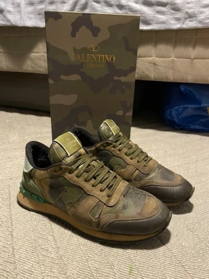 Valentino Garavani camo sneakers - Snygga sneakers från Valentino Garavani i camomönster med gröna och bruna nyanser. Skorna har klassisk låg modell, gröna skosnören och detaljer i läder och textil. Bakpartiet har en cool nitar-detalj i grönt. Perfekta för dig som gillar streetstyle och exklusiva märken.