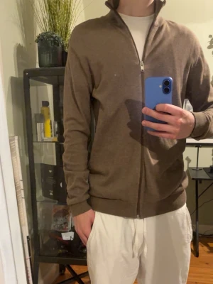 Selected homme tröja - Säljer en stilren beige ziptröja med hög krage och hel dragkedja framtill. Tröjan har ribbade muddar vid ärmslut och nederkant, och är tillverkad i ett mjukt, tunt material som passar perfekt till lager-på-lager. Enkel och clean design. Orginal pris cirka 450 kr